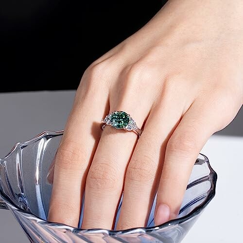 Hand wearing a silver ring with a green gemstone over a decorative bowl.